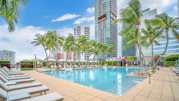 A swimming pool with lounge chairs, palm trees, and modern buildings in the background.