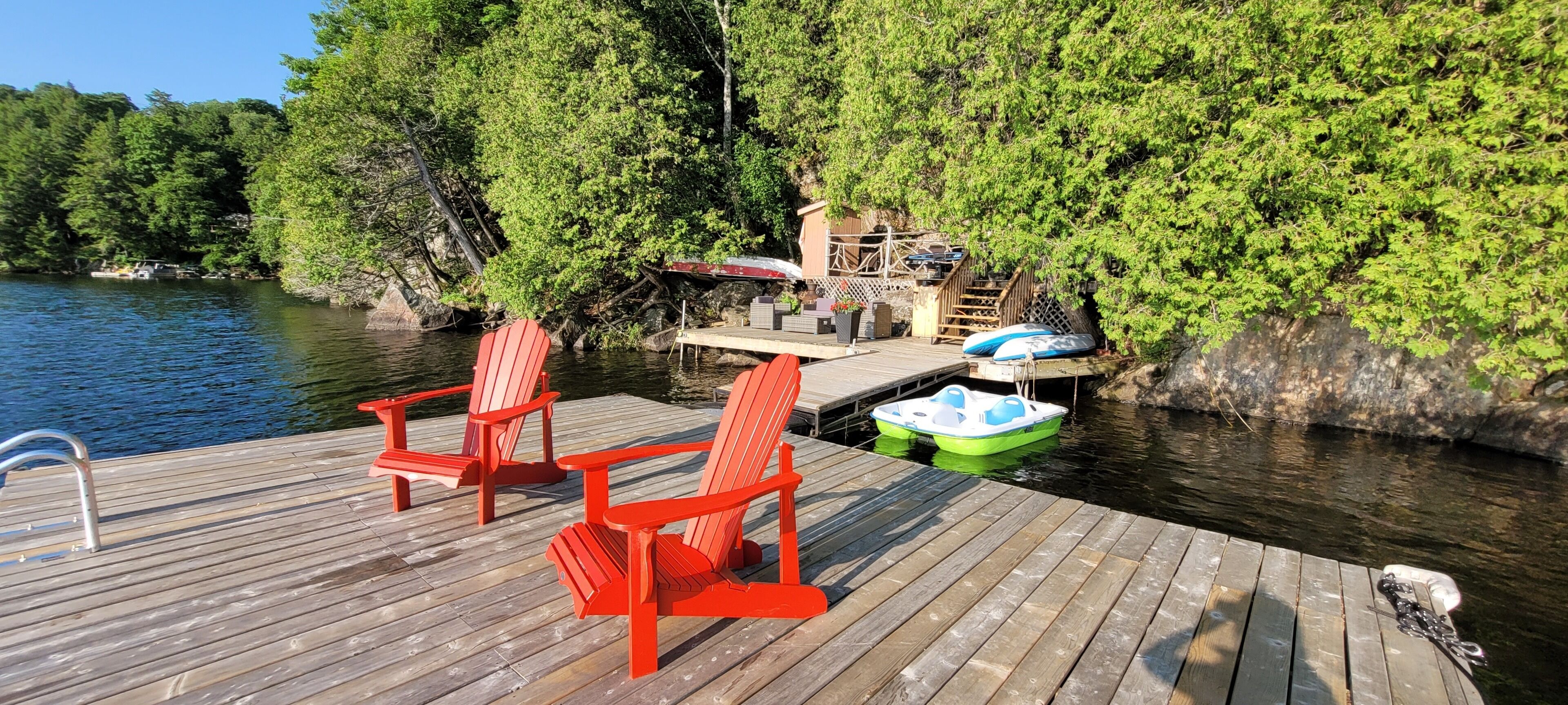 Une jetée en bois avec des chaises Adirondack rouges, un bateau vert et blanc, et un petit bateau amarré.