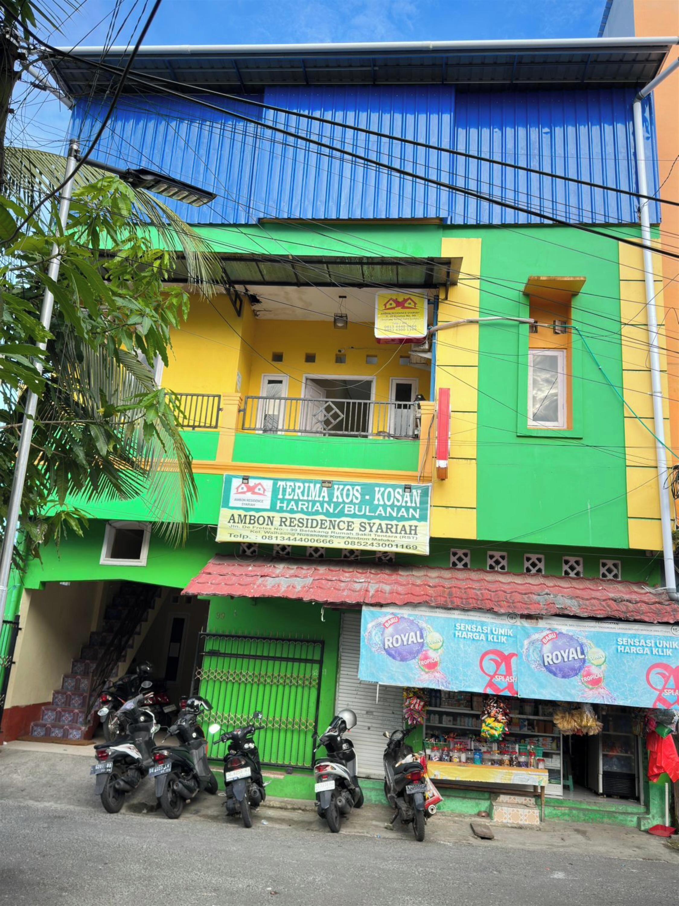Un edificio colorido con un cartel que dice 'Terima Kos. Kosan Harian/Bulanan' y 'Ambon Residence Syariah'. Hay motocicletas estacionadas en la parte delantera.