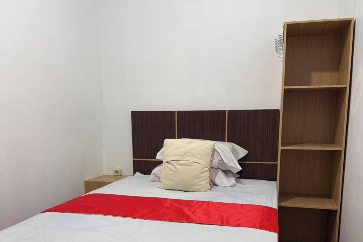A single bed with a white mattress and a red bedspread, a wooden headboard, a small bedside table, and a wooden wardrobe.