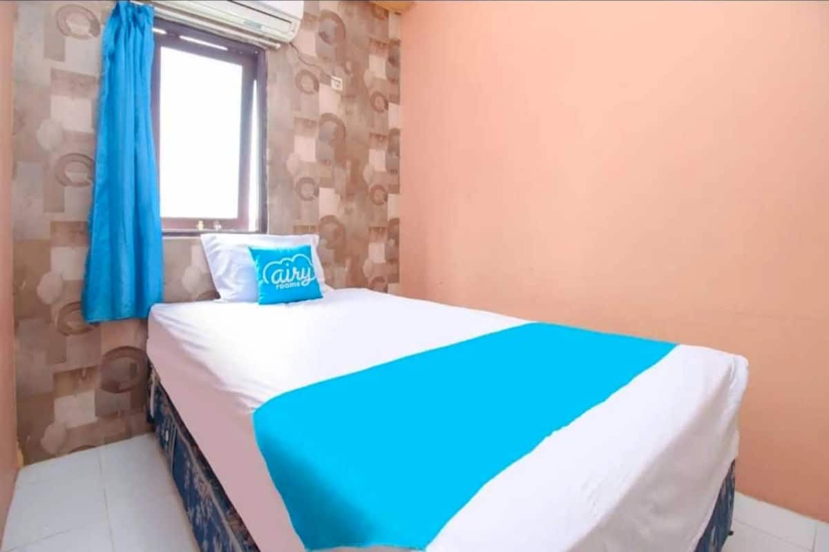 A single bed with a blue and white bedspread, a pillow with the text "Cairu Room," and a window with blue curtains.
