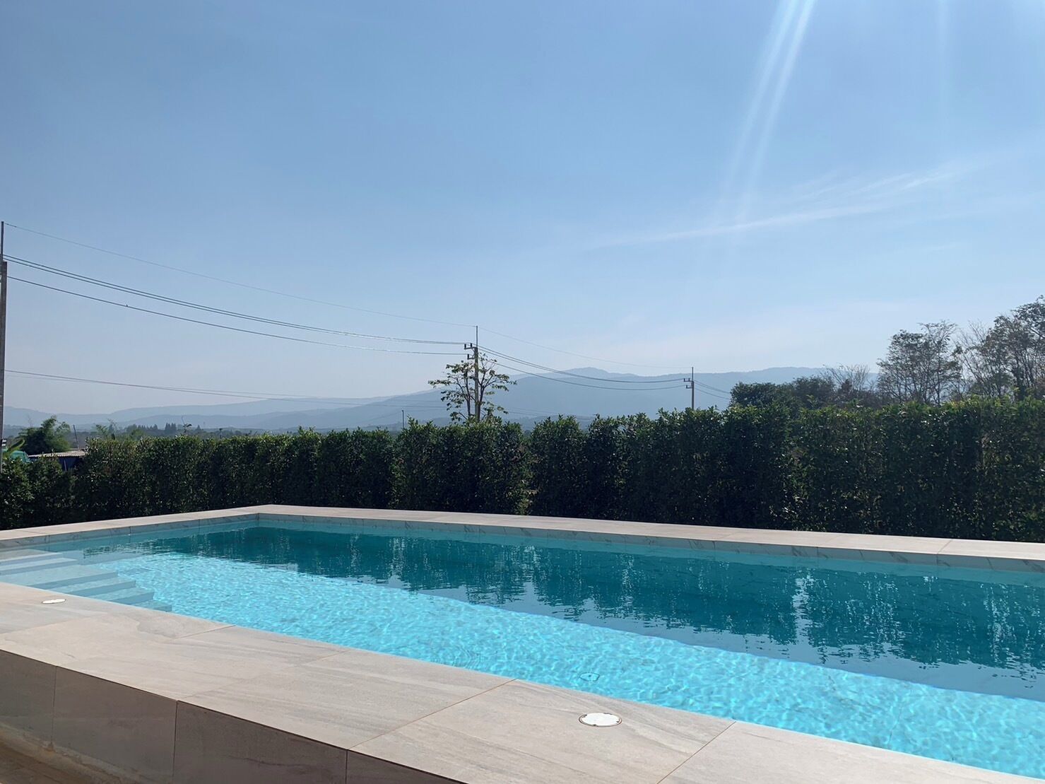 Une piscine rectangulaire à l’eau d’un bleu limpide, entourée d’une terrasse carrelée. Au fond, on aperçoit des arbustes verts et un ciel dégagé avec des montagnes lointaines.