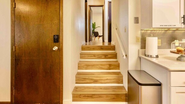 A wooden staircase leading to a room with a kitchen area, including a countertop with a paper towel dispenser and a trash can. - Blue House Retreat 2 (Sioux Falls)