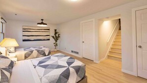 A bedroom with two beds, a nightstand, a lamp, a ceiling light, a wall art piece, and a staircase leading to another room. - Blue House Retreat 2 (Sioux Falls)