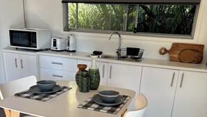 A modern kitchen with white cabinets, a dining table with two chairs, and a window with a view of greenery. - Studio 59 - Byron Hinterland. (Myocum)