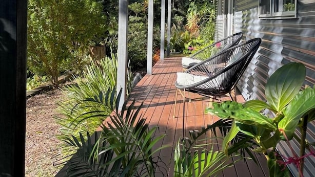 A covered outdoor deck with a wooden floor, surrounded by lush greenery and a house with large windows. - Studio 59 - Byron Hinterland. (Myocum)