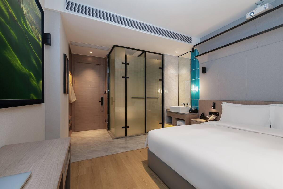 A modern hotel room with a large bed, a bedside table, a built-in desk, a glass-enclosed walk-in closet, a bathroom with a mirror and sink, and a large TV screen.