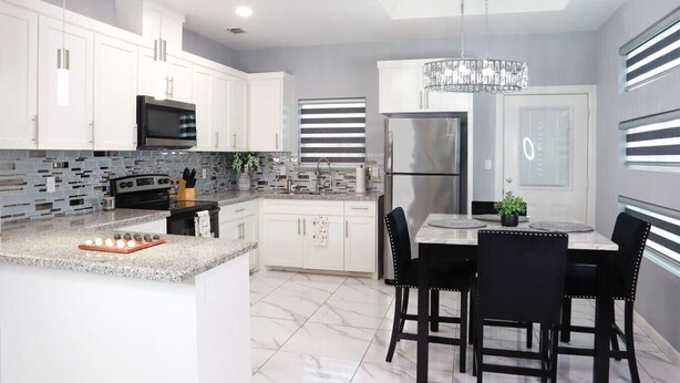 A modern kitchen with white cabinets, a stainless steel refrigerator, a built-in oven, and a dining area with a marble countertop and black chairs.