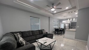 A modern living room with a sectional sofa, a marble coffee table, and a dining area with a kitchen in the background.