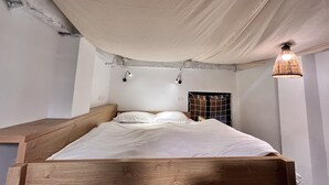 A bedroom with a wooden bed, white bedding, a hanging light fixture, and a wall-mounted shelf.