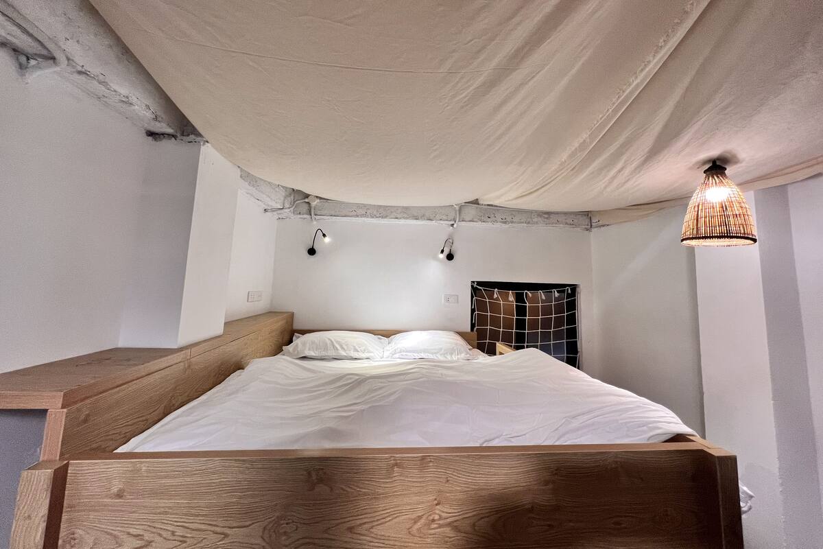 A bedroom with a wooden bed, white bedding, a hanging light fixture, and a wall-mounted shelf.