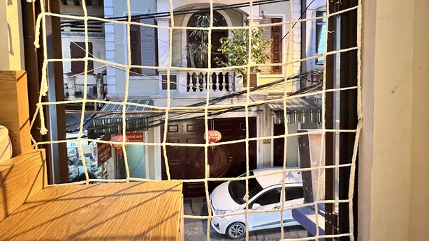 A view through a window with a wooden frame and a white net barrier. A white car is parked outside, and a building with balconies and a door is visible in the background.