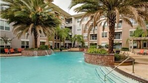 A swimming pool with palm trees and lounge chairs, surrounded by a multi-story building with balconies.