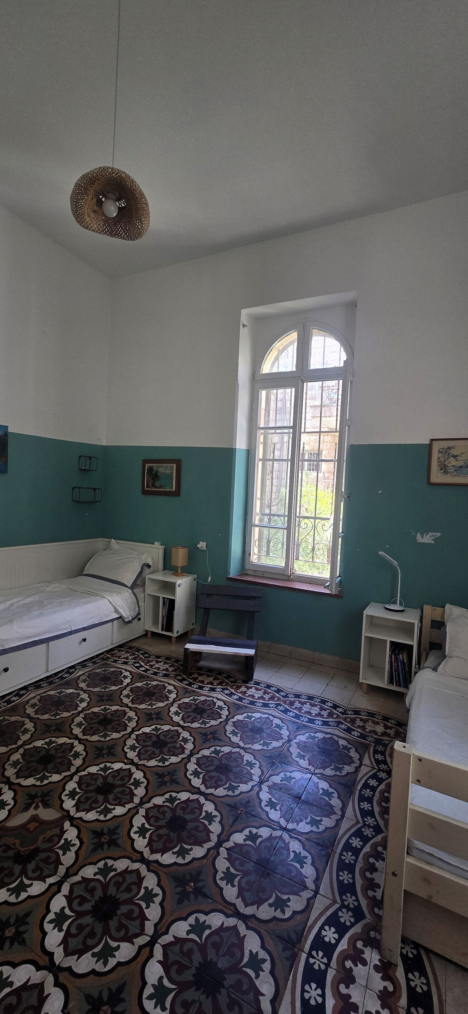 Ein Zimmer mit einem Bett, einem Schreibtisch, einem Stuhl, einem Fenster und einem gemusterten Boden.