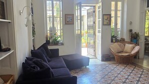 A spacious living room with a dark-colored sofa, a small wooden table, and a patterned floor. There are two large windows with decorative glass panes, and a wicker chair is visible in the background. - Historical apartment in the hart of Jerusalem (Jerusalem)