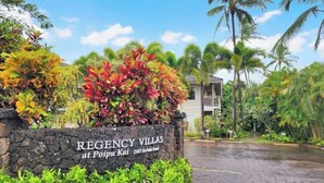 Schild für Regency Villas in Poipu Kai, zeigt eine Steinmauer mit Name und Adresse, umgeben von üppiger tropischer Vegetation und Palmen.