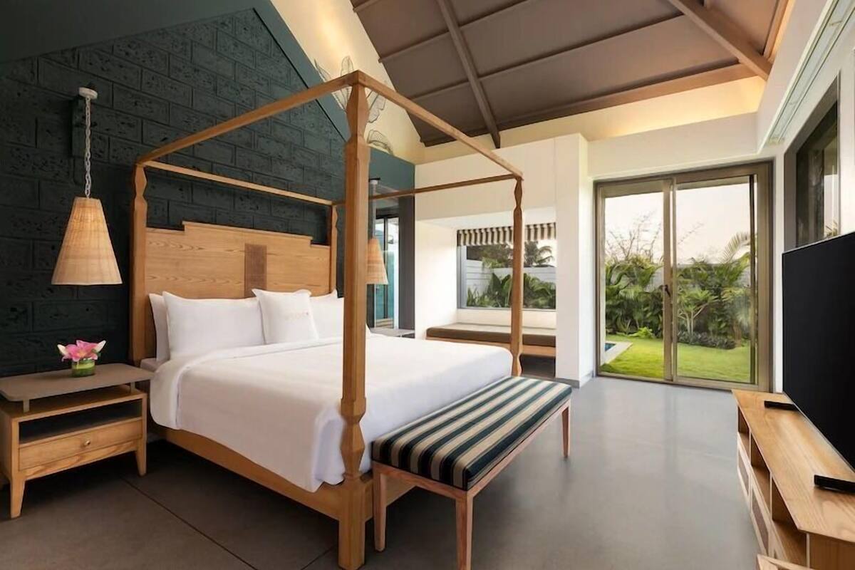 A four-poster bed with a wooden frame and white bedding, a bedside table with a lamp, and a large window offering a view of greenery.