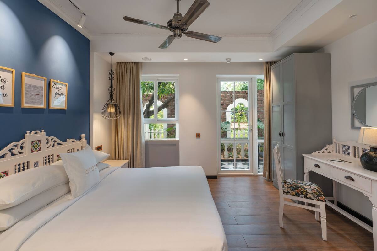 A bedroom with a large bed, a desk, and a chair. There is a ceiling fan, a window with curtains, and a view of the outside.