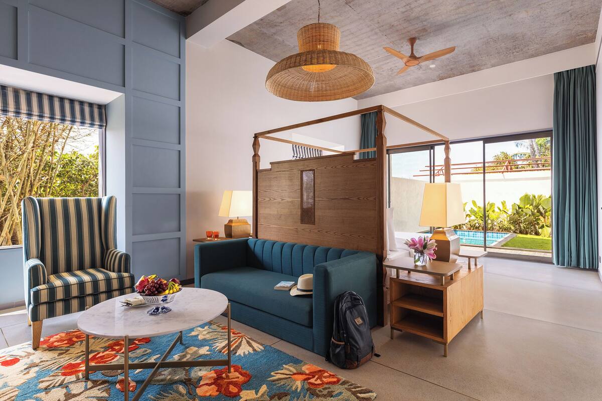 A modern hotel room with a blue sofa, striped armchair, and a wooden desk with a lamp.