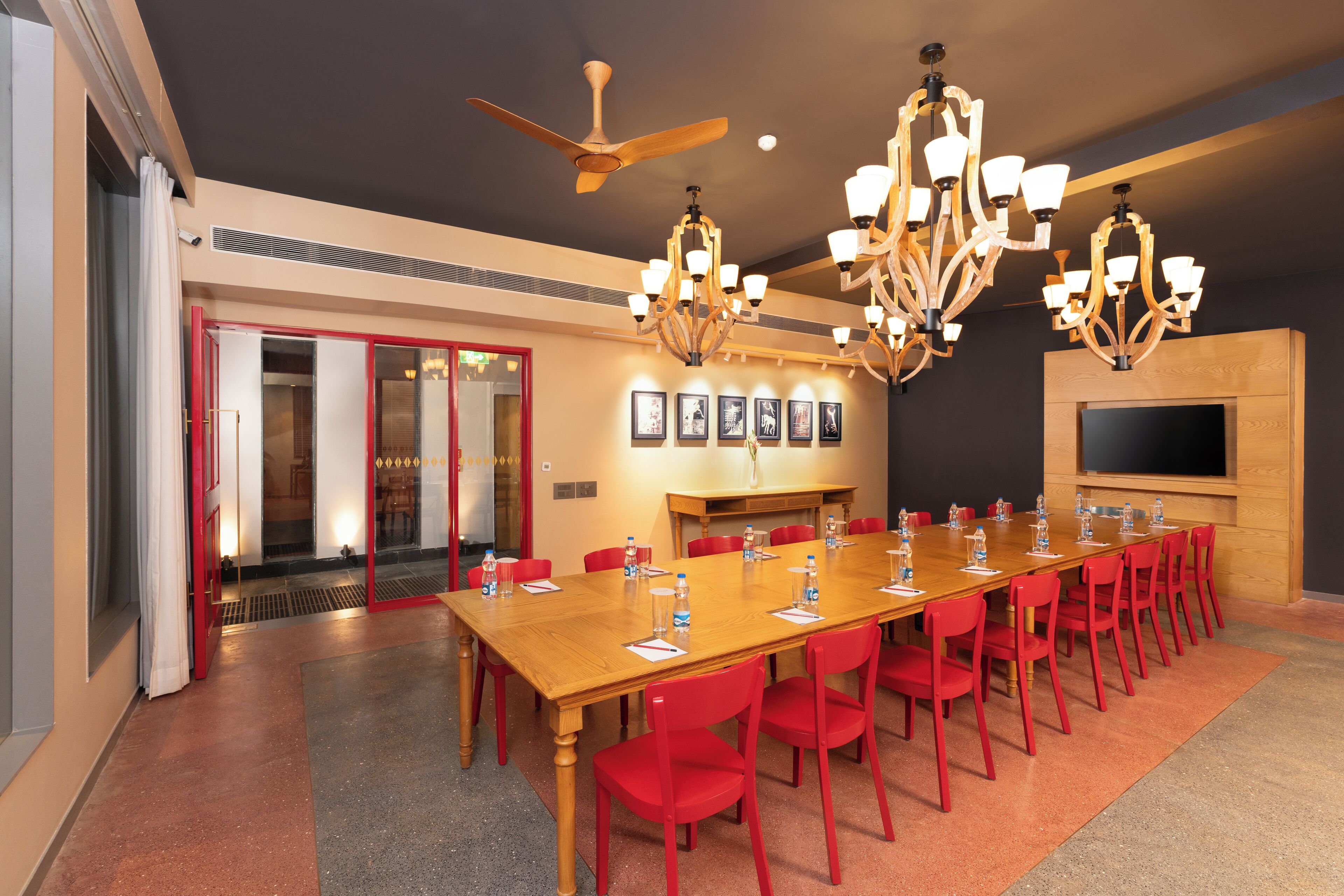 A conference room with a long table, red chairs, chandeliers, a ceiling fan, and a TV on the wall.
