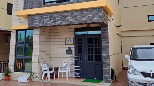 Um edifício de dois andares com varanda, um carro estacionado na frente e área de estar externa.