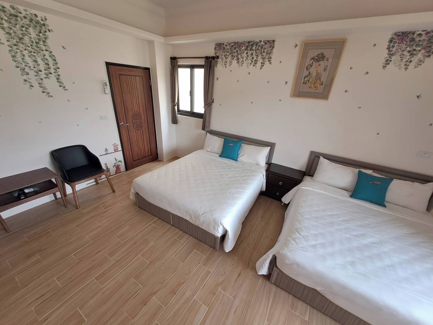 A hotel room with two beds, a desk, a chair, a wardrobe, and decorative wall art.