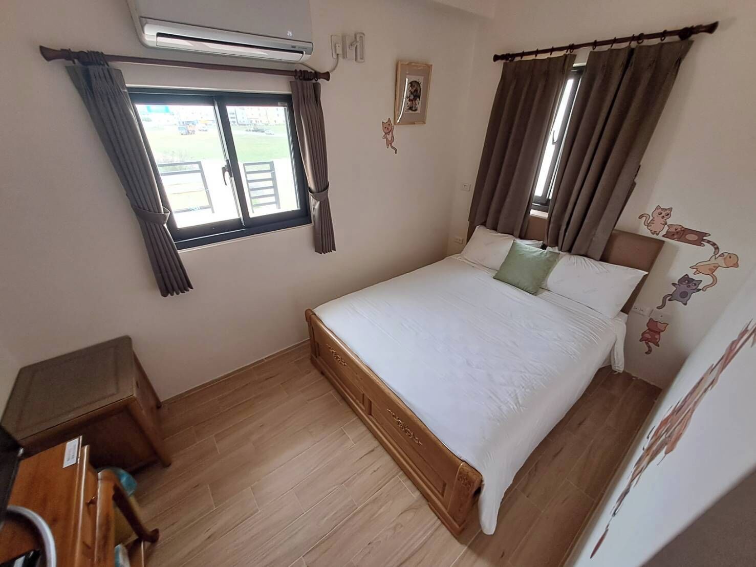 A bedroom with a bed, curtains, an air conditioning unit, a small table, and a wall with cat decorations.