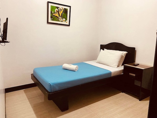 Una cama pequeña y bien hecha, con una colcha azul y una almohada blanca, una mesita de noche con un control remoto, y un cuadro en la pared.