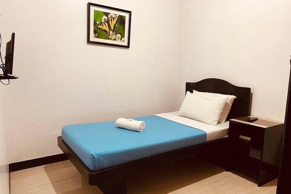 Una cama individual con colchón azul, almohada blanca y cabecera negra. Hay una mesita de noche con una lámpara y un cuadro enmarcado en la pared sobre la cama.