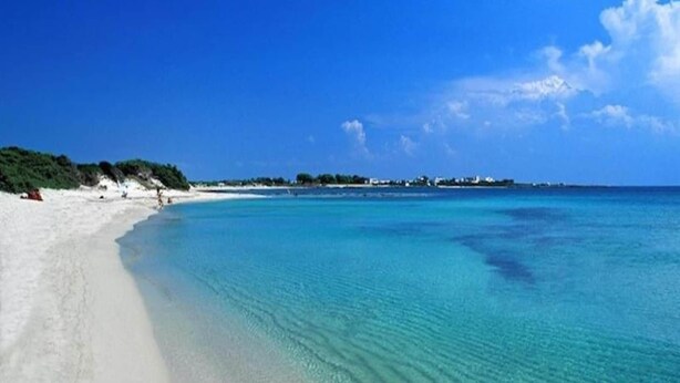 A beach with clear turquoise water, white sand, and a distant shoreline with buildings.