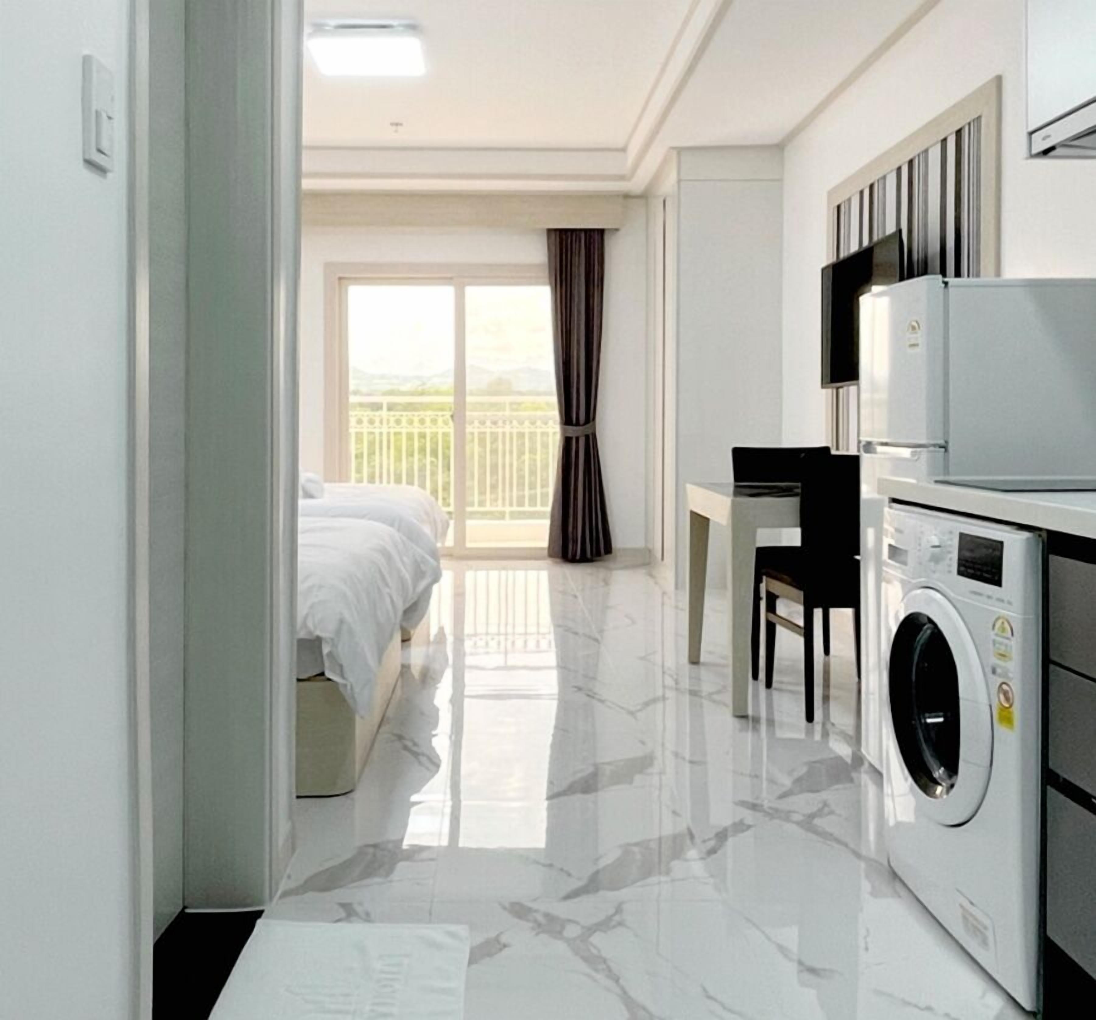 A hotel room with a bed, a desk, a washing machine, and a balcony with curtains.