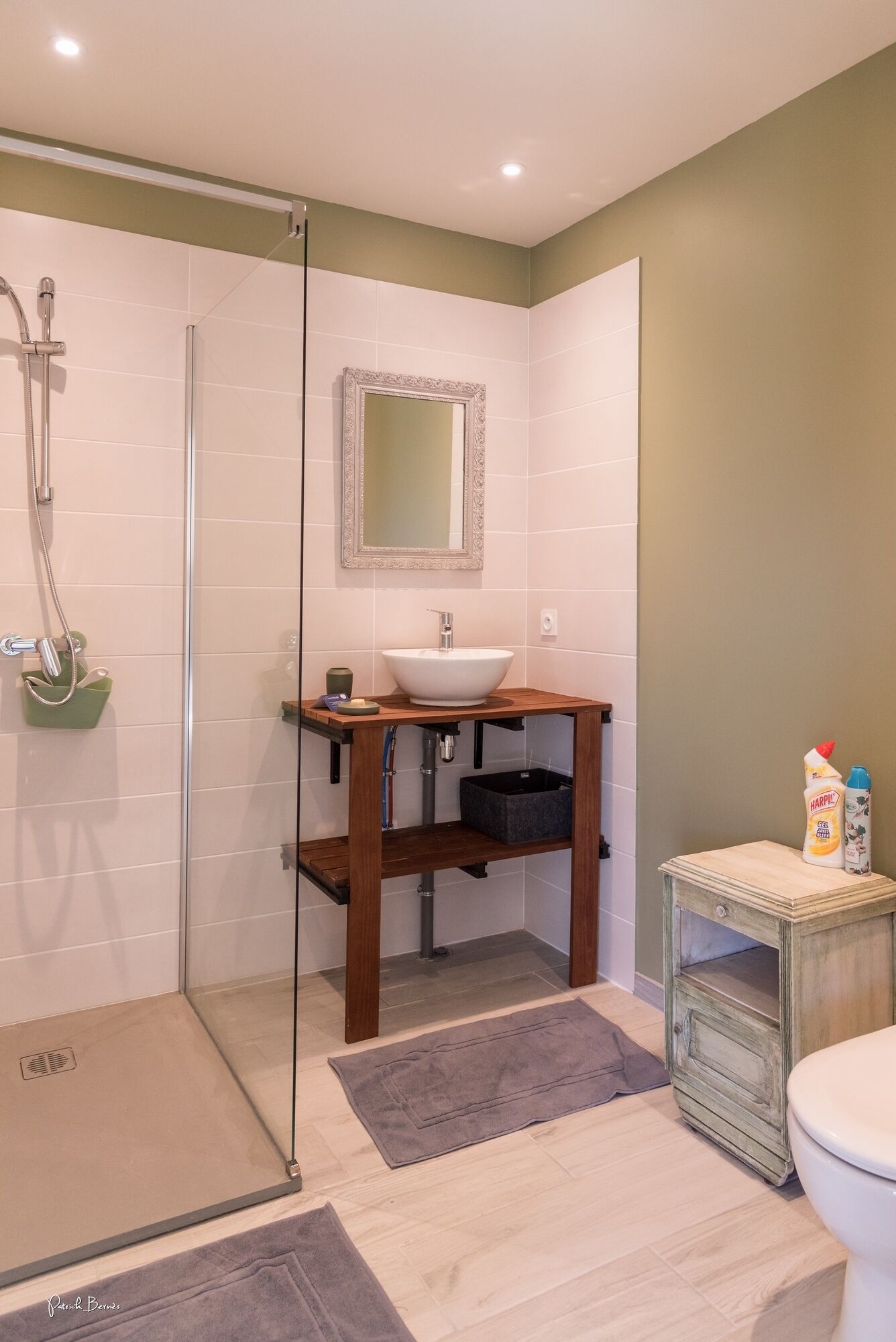 Een badkamer met een douchecabine van glas, een houten wastafelmeubel met een witte wastafel en een toilet met een klein kastje ernaast.