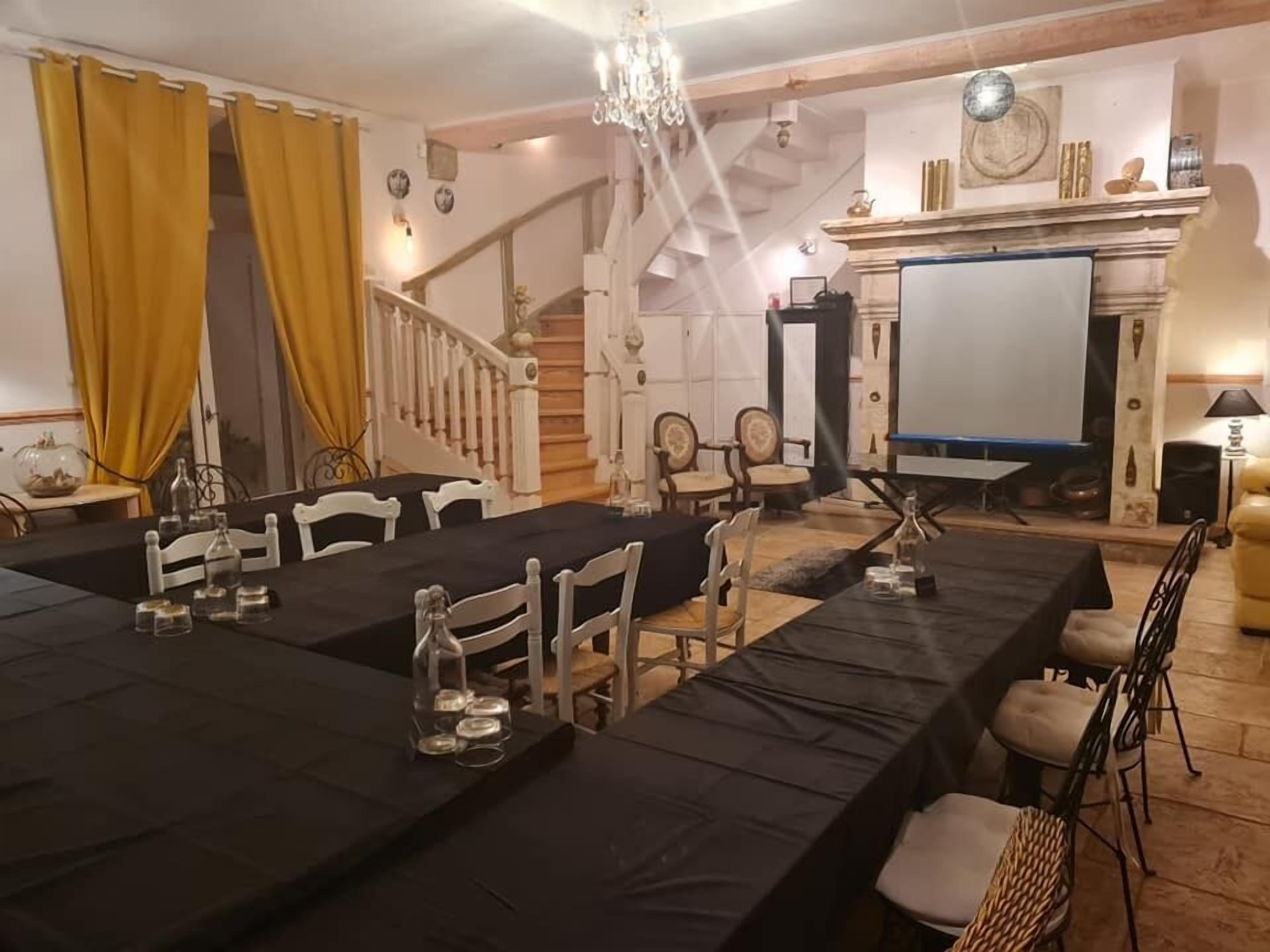 Une salle à manger avec une longue table dressée pour un repas, entourée de chaises. Un escalier avec une rampe en bois est visible au fond.