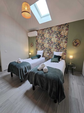 Une chambre avec deux lits, une lucarne, un papier peint à motifs floraux et une petite table avec une lampe.