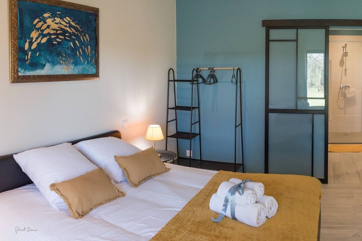 Habitació d'hotel amb un llit, una taula de nit amb una làmpada, un penjall toveles, un mirall i una zona de dutxa.