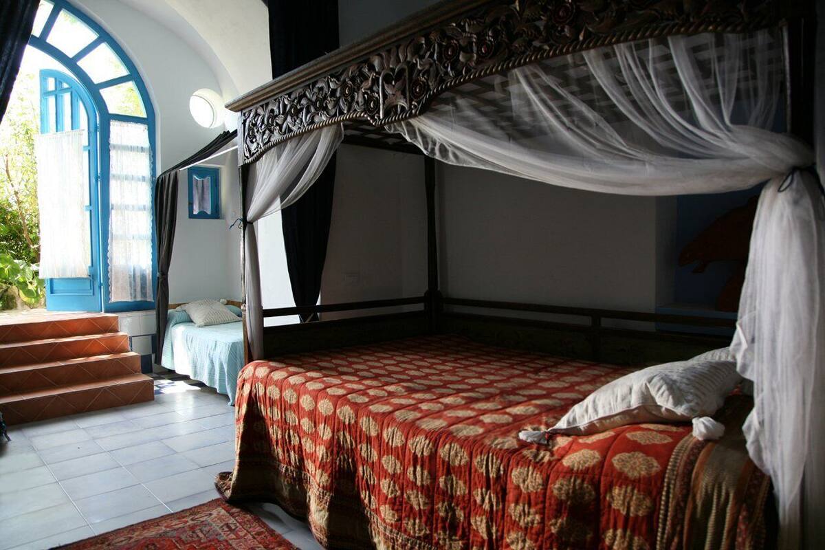 Una cama con dosel y cuatro columnas, una mesita de noche pequeña, y vistas a una puerta azul y escalones que dan al exterior.