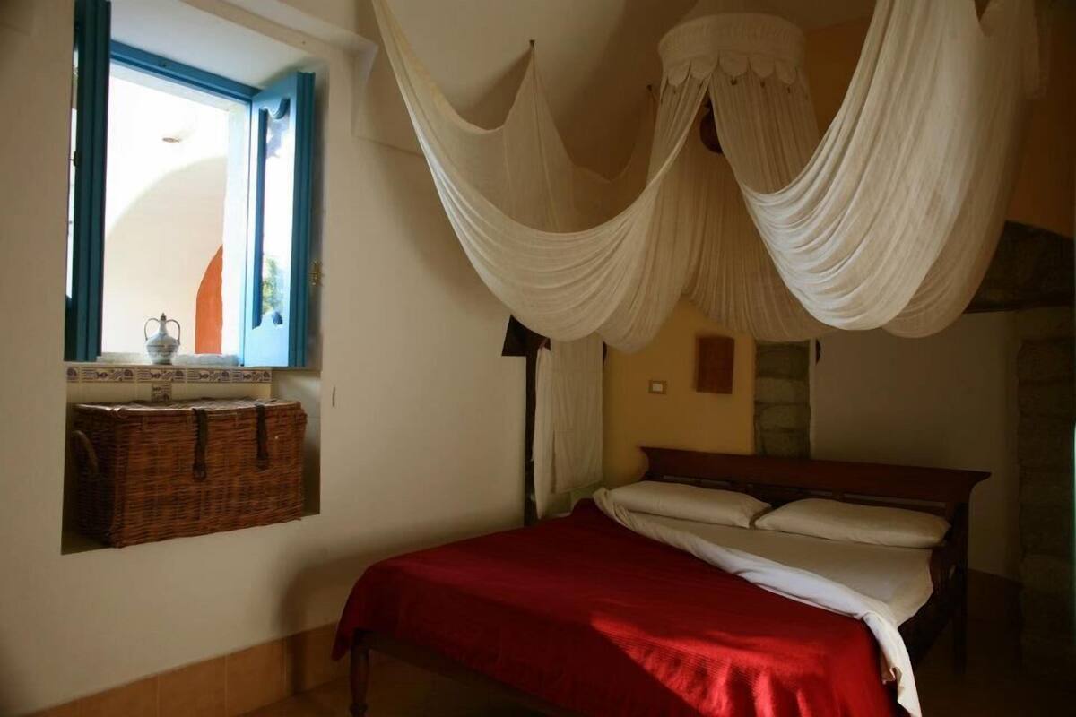 Un dormitorio con cama, una ventana con cortinas, una mesita de noche y un dosel colgante.