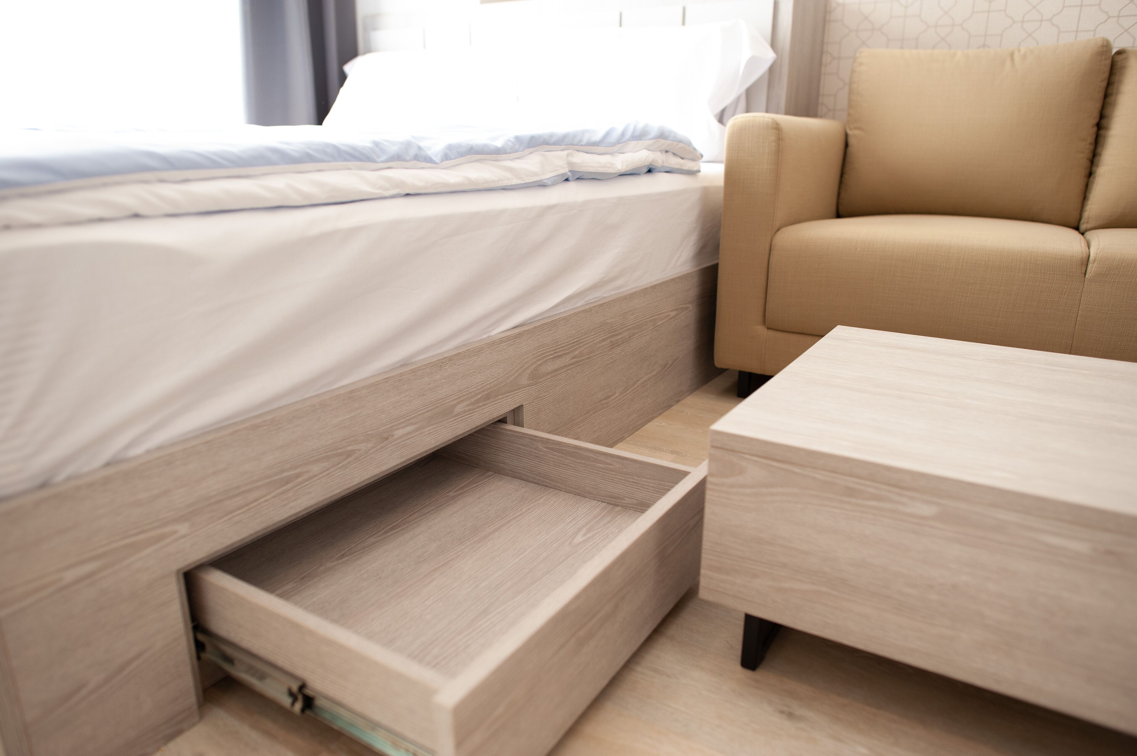 A bed with a built-in drawer, a wooden nightstand, and a beige sofa in a modern room.