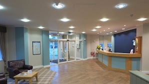 A reception area with a wooden reception desk, a waiting area with a brown leather chair and a plaid rug, and a glass door leading outside. - Burn Hall Hotel (York)