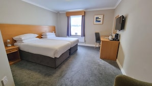 A hotel room with two beds, a desk, a chair, a TV, and a window with curtains. - Burn Hall Hotel (York)