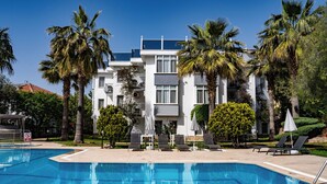 Piscina com espreguiçadeiras e guarda-sóis, cercada por palmeiras e um edifício de vários andares com varandas.