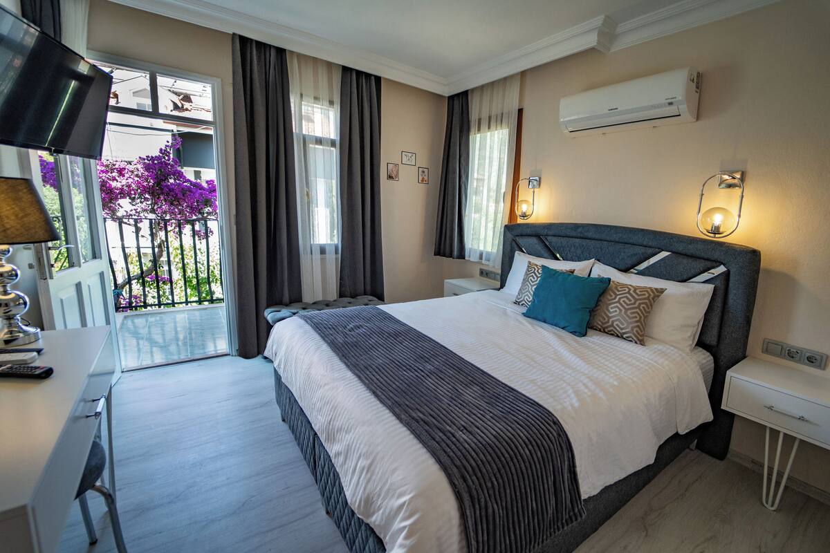 Habitación de hotel con una cama grande, un escritorio, una silla y un balcón con flores moradas.