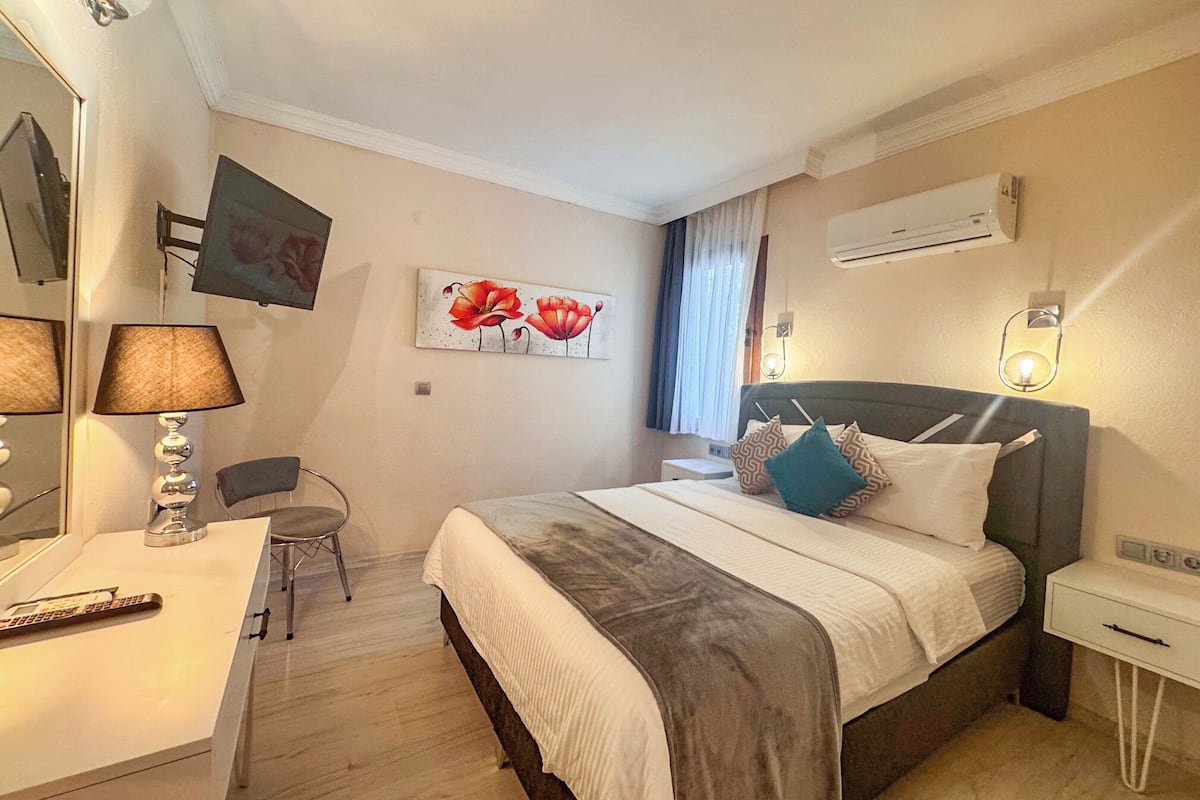Una habitación de hotel con cama, mesita de noche, lámpara, sillón y una obra de arte en la pared.