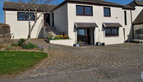 A two-story house with a paved driveway, a small garden with flowers, and a staircase leading to the entrance.