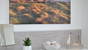 Un baño con un cuadro grande de un campo cubierto de hierba, una planta en maceta, un cuenco de conchas marinas y un barco modelo.