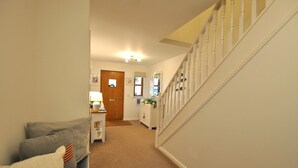 A hallway with a carpeted floor, a white staircase with white balusters, a wooden door, and a white cabinet with a lamp.