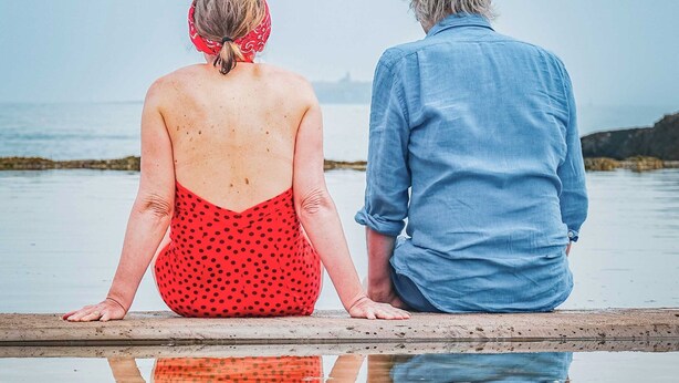 Two people sitting by a calm body of water, reflecting their image.
