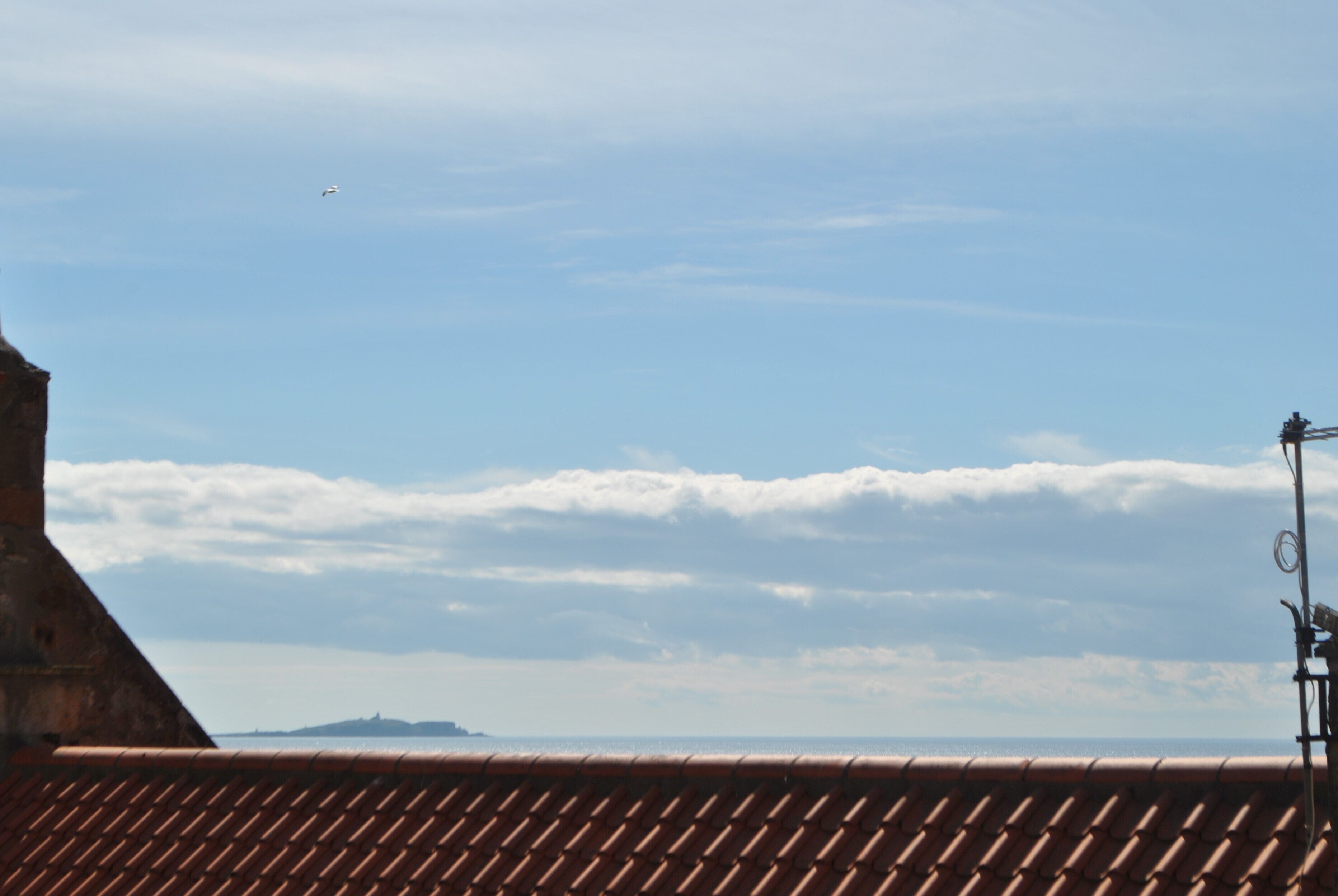 A clear sky with a plane flying overhead, a tiled roof, and a distant view of land across the water.