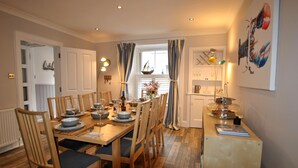 A dining area with a wooden table set for six, flanked by chairs with blue cushions. A window with curtains is visible, and a kitchen area with a counter and various items is to the right.