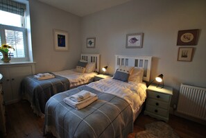 A bedroom with two beds, each with a pillow and a bedside table with a lamp. There are framed pictures on the wall and a window with blinds.